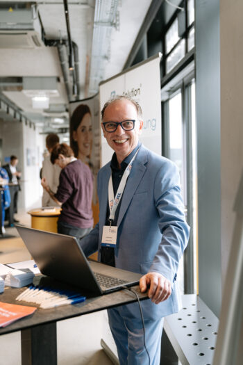 Mann im hellblauen Anzug steht mit Laptop an einem Stehtisch